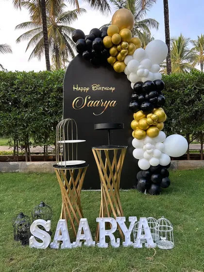 A chic outdoor birthday setup with a black, gold, and white theme. The decor features a personalized black backdrop, a stylish balloon garland, and a light-up name sign on the grass.