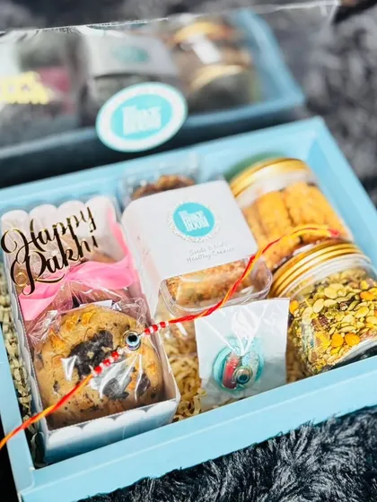 This is a sample of my thoughtfully curated Rakhi hamper. It includes a variety of treats like cookies and sweets, all beautifully packaged in a festive blue box with a 'Happy Rakhi' message and a rakhi.
