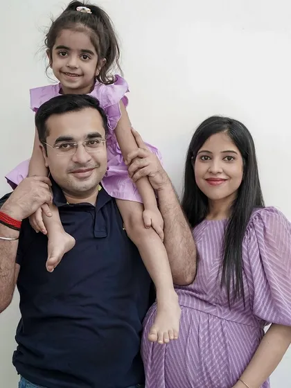 A happy family portrait celebrating their second pregnancy. The father holds their daughter on his shoulders while standing next to his expecting wife, all coordinated in lovely lavender outfits.