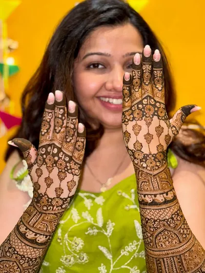 A happy bride showing off her beautiful back-of-hand mehendi. The symmetrical design features delicate hanging motifs and intricate patterns, creating a very elegant look.