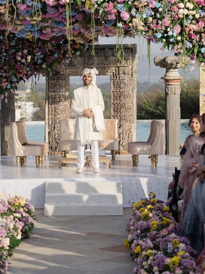 The groom awaits his bride under a stunning pastel floral mandap. The structure, combining ancient stone pillars with a modern, dense floral canopy, perfectly blends the venue's heritage with a fresh, romantic aesthetic.