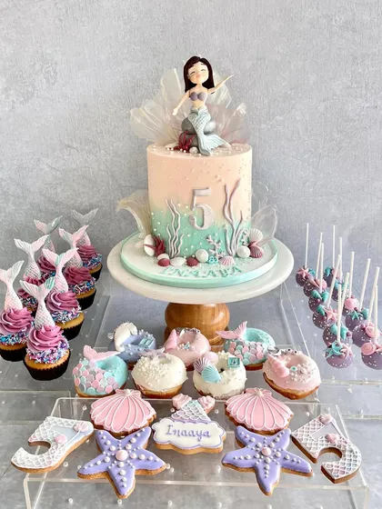 A complete mermaid dessert table for Inaaya's 5th birthday. The setup includes a beautiful cake, cupcakes with mermaid tails, and custom-decorated cookies and donuts.