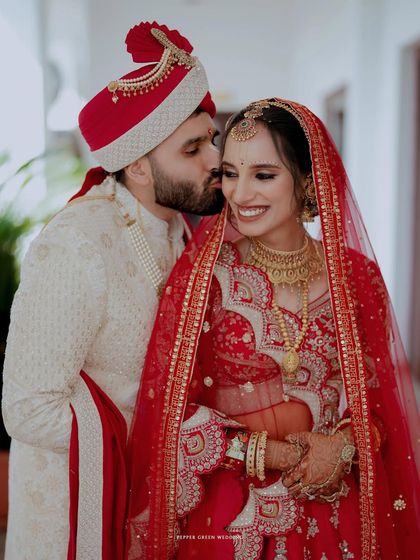 A sweet moment as the groom kisses his bride's cheek. Her joyful smile and the rich red of her lehenga make this a beautiful wedding portrait.