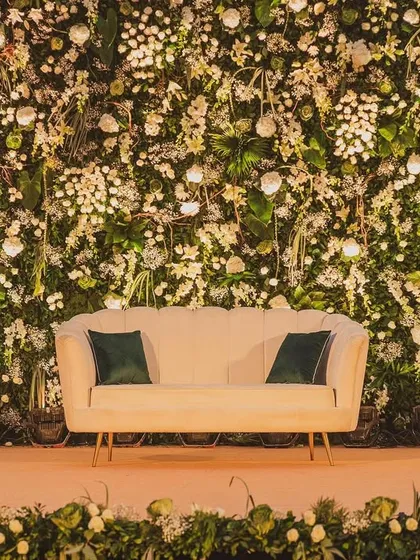 A close-up of a reception photo booth backdrop, featuring a dense wall of white flowers and lush greenery. The lighting is carefully placed to create a warm, inviting glow for perfect pictures.