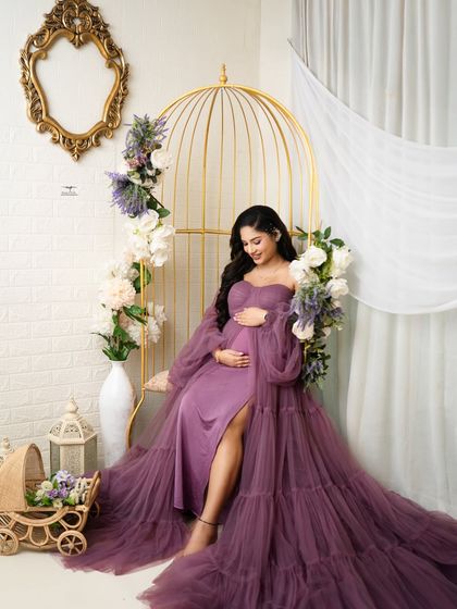 Radiant in a flowing purple gown. The mother-to-be cradles her bump while seated in the golden birdcage chair, surrounded by soft lavender and white flowers.