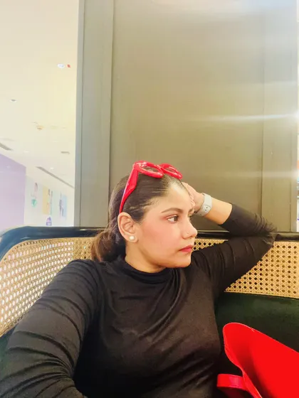 A candid shot while seated, wearing a simple black top and red heart-shaped glasses, showing a relaxed yet stylish moment.