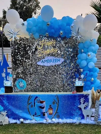 A magical Frozen-themed birthday party setup. The centerpiece is a shimmering silver sequin backdrop, framed by a blue and white balloon garland, with cutouts of Elsa's castle and other winter elements to create an enchanting wonderland.