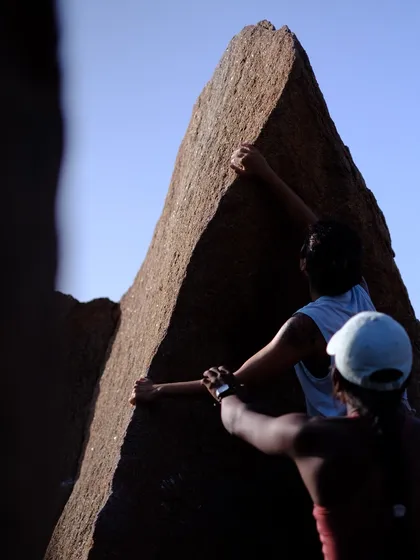 The art of spotting is crucial in bouldering. Here you can see the spotter's hands ready to guide and protect the climber on the sharp arete of this Hampi boulder.