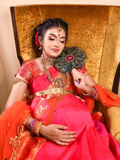 Royalty and tradition combined. This stunning portrait features a mom-to-be in an ornate pink and orange silk saree, holding a peacock feather fan on a golden throne.