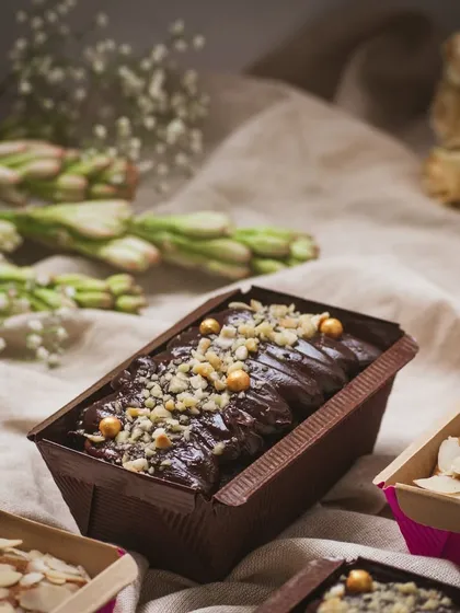 A close-up on a gluten-free chocolate loaf cake, garnished with nuts. The focus is on the rich texture and glossy ganache, making it look incredibly appetizing.