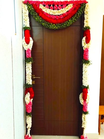 A simple door decoration for a housewarming ceremony. The door is framed with garlands of jasmine, rose petals, and other colorful flowers.