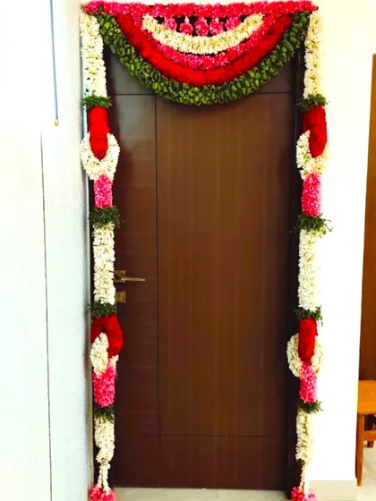 A simple door decoration for a housewarming ceremony. The door is framed with garlands of jasmine, rose petals, and other colorful flowers.