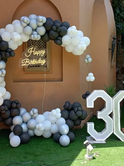 An outdoor 30th birthday party with a monochrome theme. The setup includes a circular balloon arch in black, white, and grey, a neon sign, and large '30' marquee numbers.