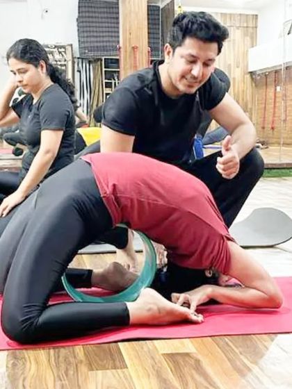 A teacher guides a student in Kapotasana (Pigeon Pose) using a yoga wheel, ensuring a safe and deep opening of the shoulders and thoracic spine.