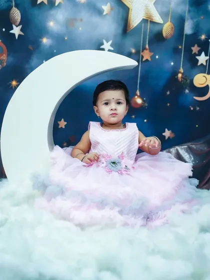 A princess on the moon. This dreamy indoor setup features a large moon prop, fluffy clouds, and a starry night backdrop for a magical portrait.