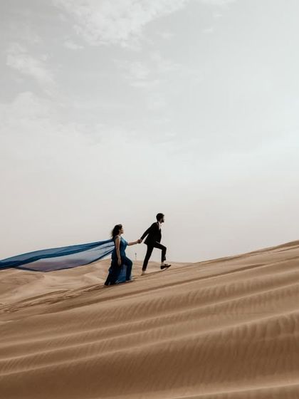 A dramatic shot of the couple walking up a sand dune, with the bride's long blue fabric trailing behind her.
