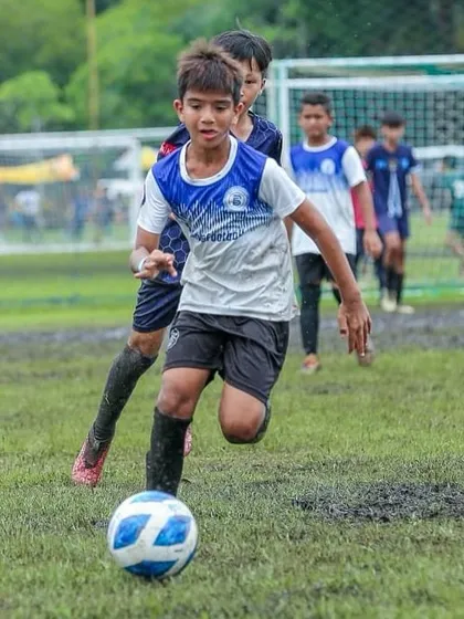 A U11 player navigates a muddy pitch with determination, showcasing his skill and power. This action shot celebrates a player who is a true team player, willing to do whatever it takes.