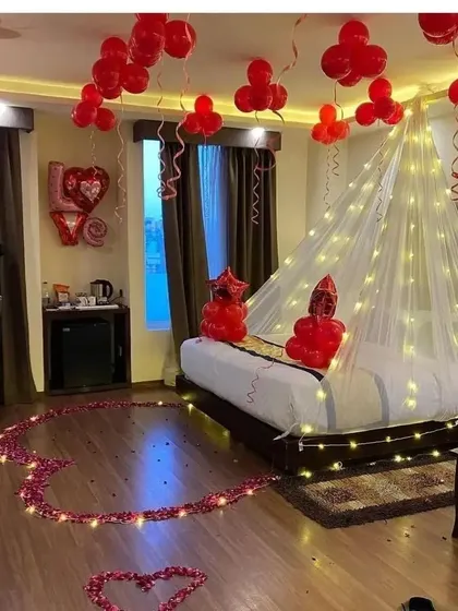 A romantic hotel room decoration with a heart made of rose petals on the floor, a light-up canopy over the bed, and red balloons.