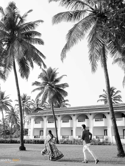 A dynamic black and white shot of the couple running through a grove of palm trees, capturing their playful energy and the iconic Goan landscape.