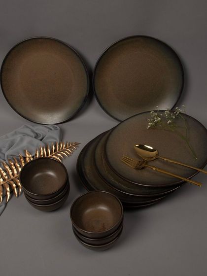 A full dinnerware set in a dark, rustic finish, styled with a golden leaf and grey fabric for a premium look.