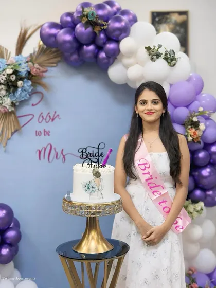 The bride-to-be posing with her beautiful cake. The backdrop features a balloon arch in shades of lavender, lilac, and white, with dried floral accents for a touch of rustic charm.