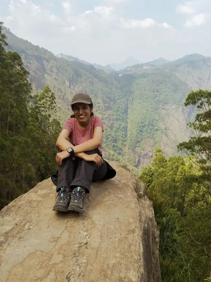 A woman sits happily on a rock, with the vast green valleys of Kodaikanal behind her. This is a moment of pure joy and connection with nature.