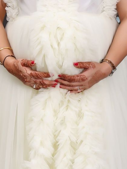 A close-up shot focusing on the details: henna-adorned hands with red nail polish gently holding a baby bump covered by a white ruffled gown. A beautiful detail of the pregnancy journey.