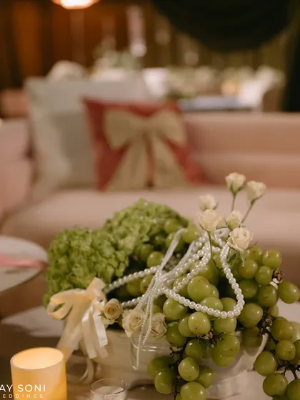 A centerpiece detail combining green grapes, hydrangeas, and delicate pearls, with a bow-themed cushion in the background, perfectly encapsulating the event's theme.