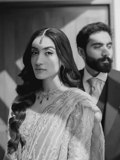 An artistic and moody black and white portrait. The focus on the bride's direct gaze, with the groom a soft presence behind her, creates a compelling and modern couple's shot.