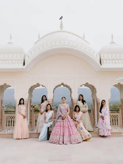 A classic group portrait of the bride with her bridesmaids, all dressed in pastel outfits, posing on a palace terrace.