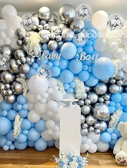 A classic 'Baby Boy' baby shower wall decoration. The dense balloon wall in shades of blue, silver chrome, and white creates a stunning backdrop, complete with a teddy bear and a pedestal for gifts or a cake.