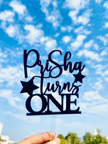 The "Prisha turns ONE" topper held against the sky, creating a striking silhouette that emphasizes its shape and design.