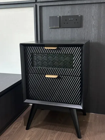 A modern black nightstand with a textured, diagonal-grooved drawer front. The small brass handle adds a touch of elegance to this contemporary piece.