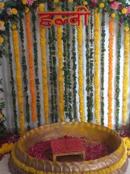 A classic Haldi ceremony arch with a mix of colorful flowers and hanging garlands. The large brass urli in the center is filled with rose petals, ready for the rituals.