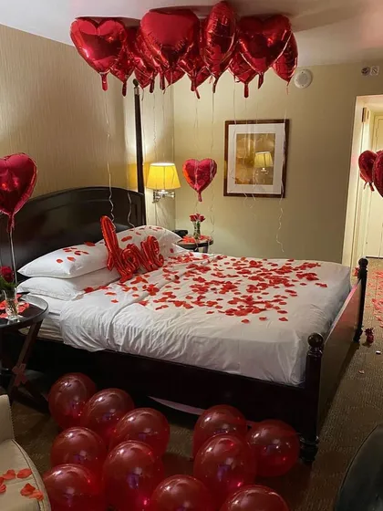 A classic romantic hotel room decoration with a bed covered in rose petals, a "Love" sign, and bouquets of red heart balloons.