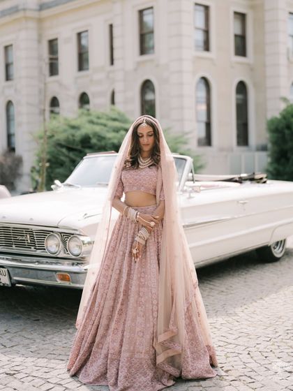 A classic bridal portrait in front of a vintage car in Turkey. The bride's soft pink lehenga and demure pose create a timeless and romantic image.