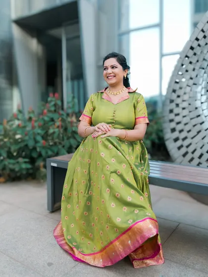 A lovely portrait of a woman in a unique green traditional dress, seated on a bench in a modern, urban setting.