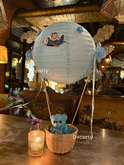 A charming table centerpiece featuring a small blue teddy bear in a hot air balloon basket. The soft candlelight adds a warm and cozy touch to the decor.