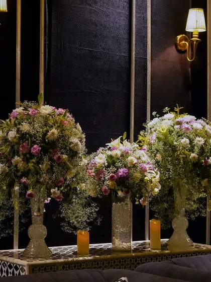 Three large floral arrangements in tall glass vases create a beautiful, symmetrical display against a paneled black wall with classic sconces.