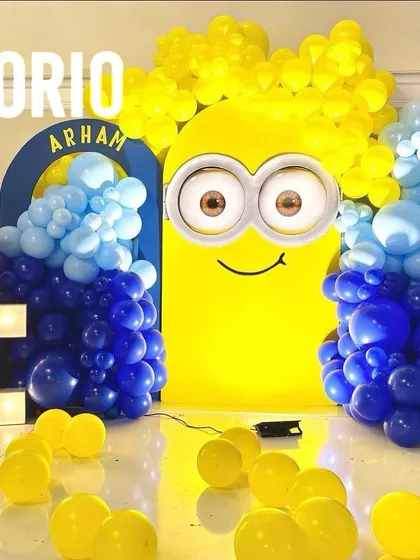 A fun and bold Minions-themed 4th birthday party. The centerpiece is a large, illuminated Minion face backdrop, surrounded by blue and yellow balloons and a '4' marquee number.