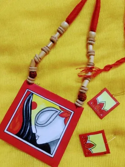 The 'Maa' Durga necklace set displayed on a bright yellow fabric, which makes the red and white painting pop. This angle shows the wooden beads on the adjustable cord.