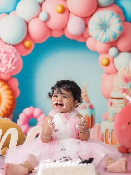 The pure joy of a first birthday cakesmash. This little girl's happy, messy smile is captured against a vibrant candy-themed backdrop with pink and blue balloons, showing a fully styled milestone session.
