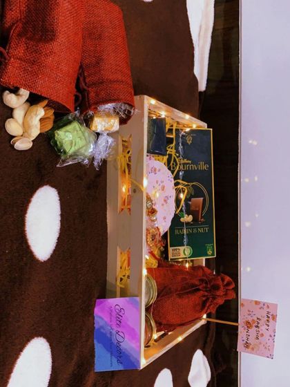 This side view shows the depth and contents of the wooden Rakhi hamper, including dry fruits spilling from a rustic jute bag, a large chocolate bar, and the beautiful rakhi.