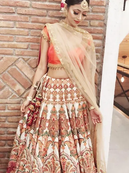 A client looking radiant in a designer lehenga by Rimple & Harpreet. The outfit features a vibrant printed skirt paired with a simple orange blouse and a light net dupatta, suitable for a mehendi or sangeet ceremony.