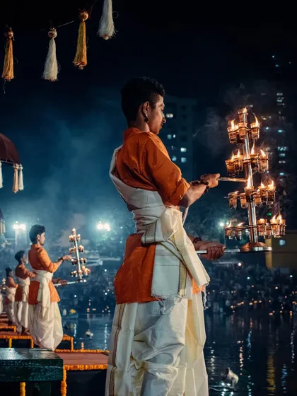 Another angle of the priest holding the ceremonial lamp, with the crowd of devotees visible in the background, highlighting the shared experience of the Dev Deepawali festival.
