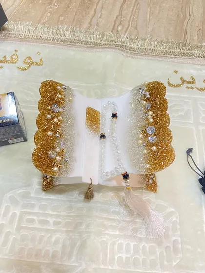 A personalized prayer set featuring a white and gold resin Quran holder with a crystal tasbeeh. The soft, light-colored prayer mat is customized with names in glittering gold script.