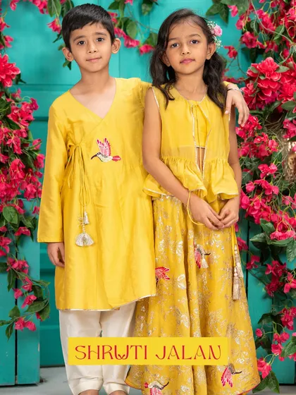 A brother and sister stand side-by-side in matching yellow outfits. His angrakha kurta features a bird motif that is also found on her floral lehenga.