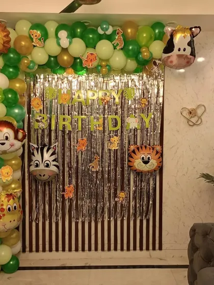A fun jungle safari theme created indoors. This decoration uses a silver fringe backdrop framed by a green, gold, and white balloon arch, complete with various animal-head foil balloons like a lion, monkey, and giraffe.