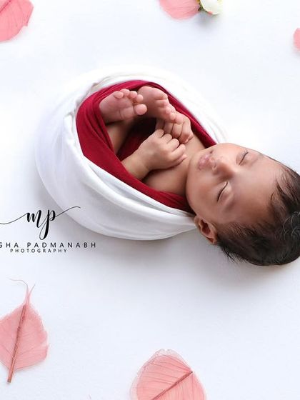 A newborn wrapped in white and red, surrounded by delicate pink leaves. The high-key white background creates a fresh and airy feel.