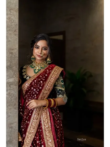 An elegant bridal portrait. The bride stands in a softly lit corridor, her luxurious red velvet saree and green jewelry creating a look of regal sophistication.